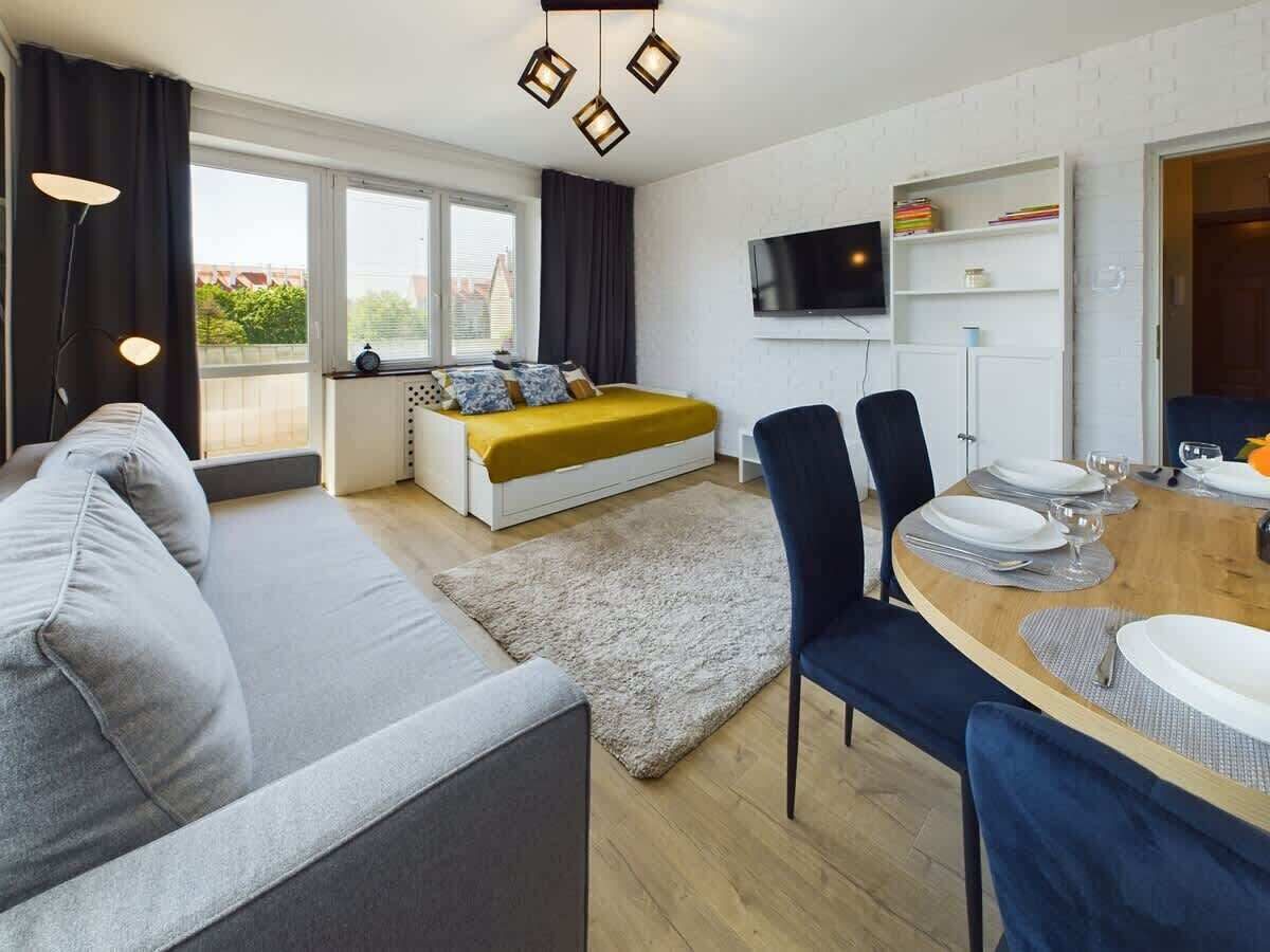 Ein modernes Wohnzimmer mit einer Couch, einem Esstisch für zwei Personen, einem Bett mit gelber Kopfteil und einem an der Wand befestigten Fernseher.