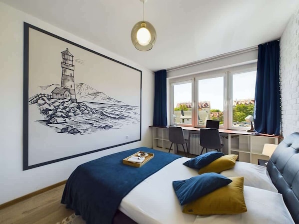 A bedroom with a bed, a desk, a chair, a large framed picture, and a window with curtains.