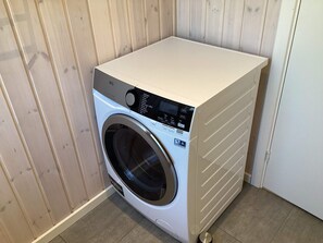 A compact, wall-mounted washing machine with a control panel, situated in a corner with wooden paneling and a tiled floor.