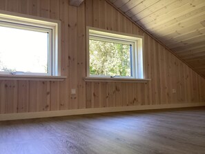 Quarto com paredes de madeira e teto inclinado, com duas janelas de moldura branca e vista para o exterior.