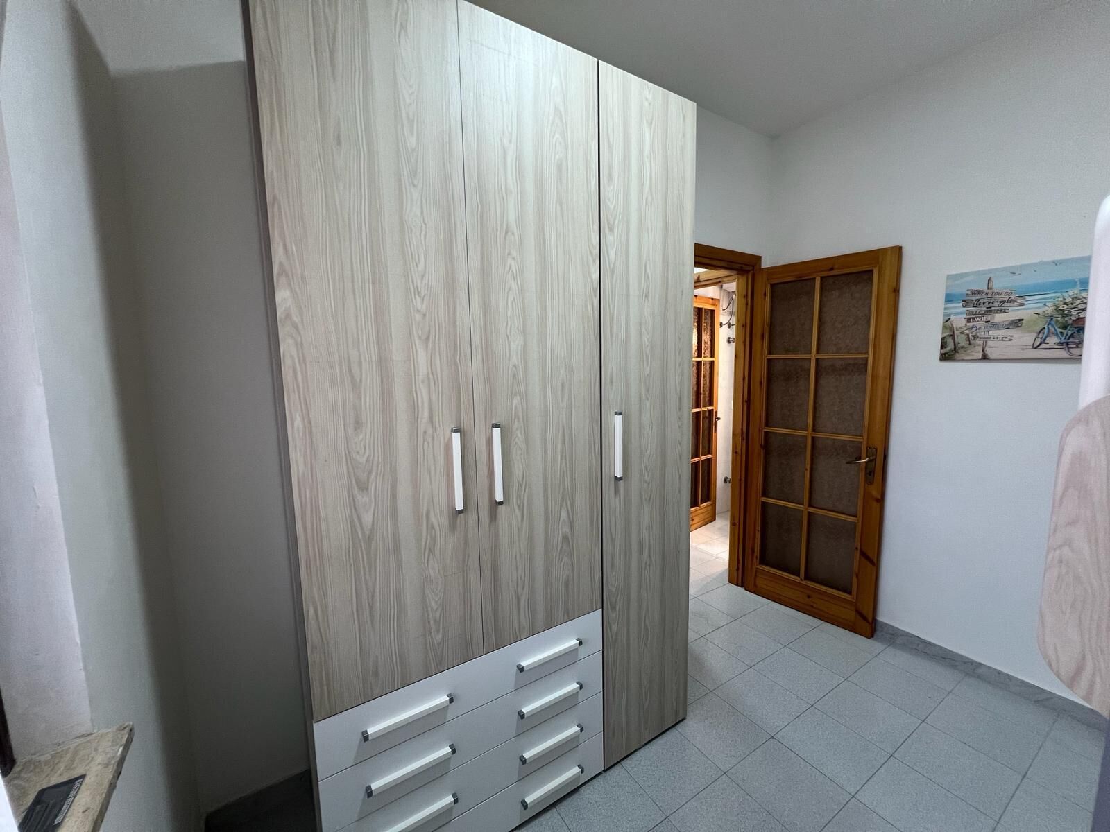 A room with a large wardrobe, a door leading to another room, and a tiled floor.