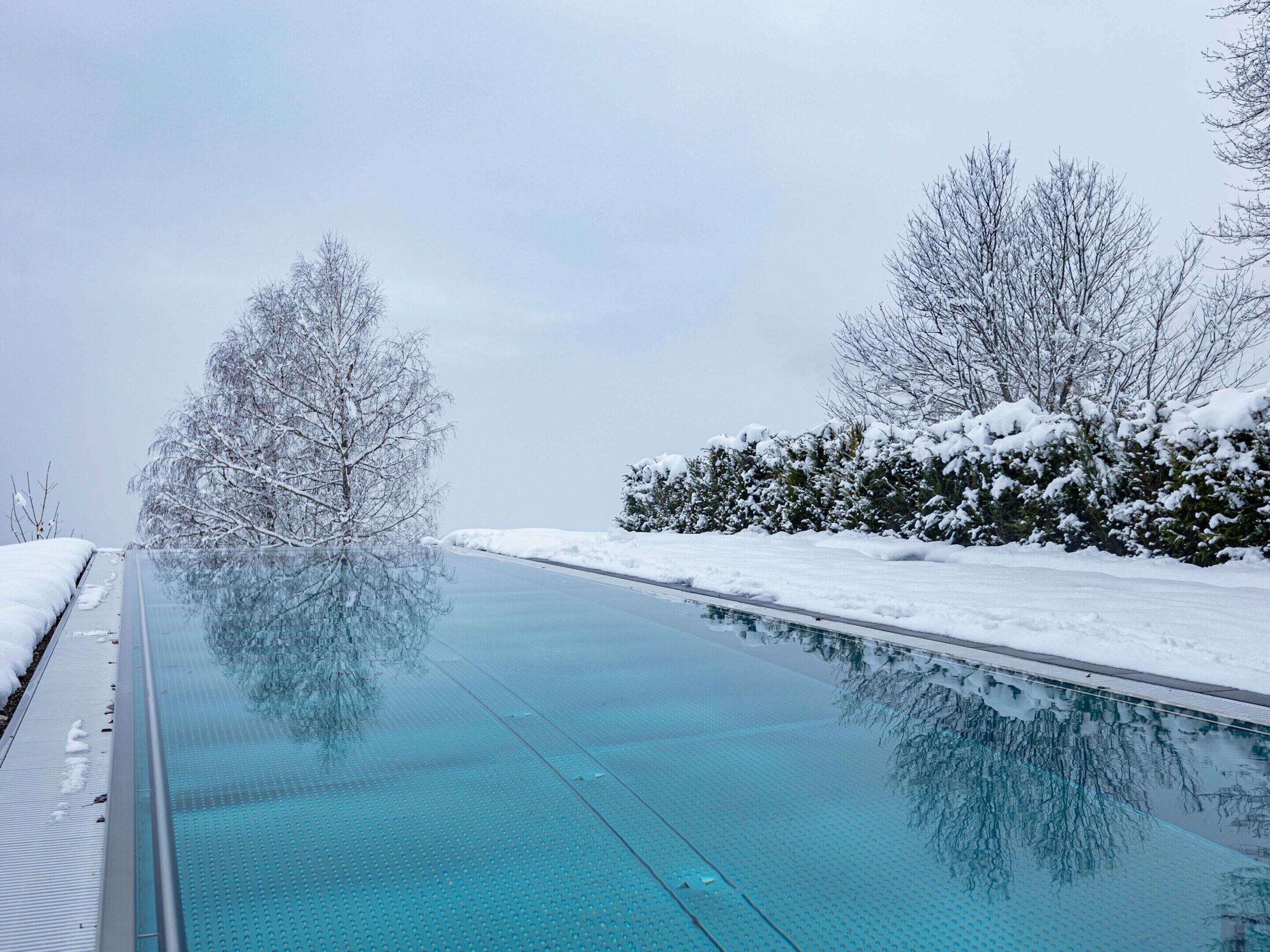 Une piscine intérieure à eau claire, séparée du reste par une paroi de verre, et entourée d’arbres et d’arbustes couverts de neige.
