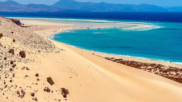 A beach with clear turquoise waters, a sandy shore, and distant mountains. - Belvilla by OYO Casa A-a3 Corralejo (La Oliva)