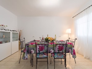 A dining area with a table covered by a floral tablecloth, surrounded by four chairs with floral cushions. A cabinet with glass doors is visible to the left. - Belvilla by OYO Casa A-a3 Corralejo (La Oliva)