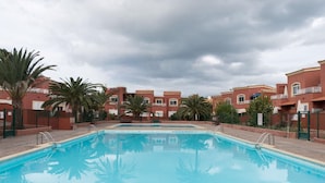 A swimming pool surrounded by red buildings with balconies, palm trees, and a cloudy sky. - Belvilla by OYO Casa A-a3 Corralejo (La Oliva)