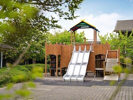 A playground with a slide and climbing area, surrounded by trees and a building with a tiled roof.