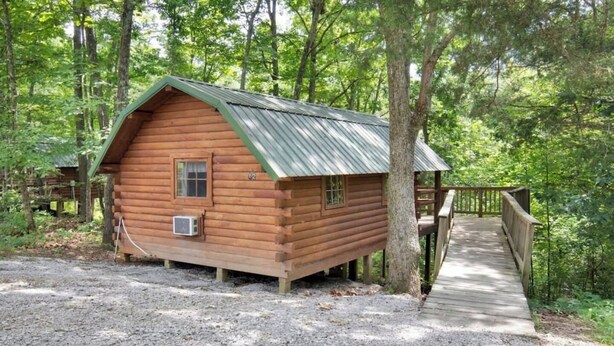 A wooden cabin with a metal roof, a small window, and an air conditioning unit. It is surrounded by trees and has a wooden deck leading to it. - Rustic Cabin in Bloomington with Views (Bloomington)