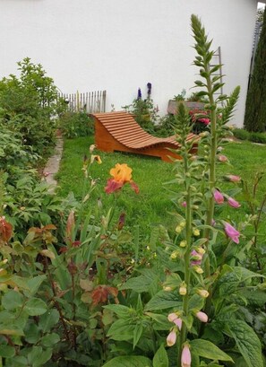 Ein Garten mit einer Holzbank, verschiedenen Pflanzen und einer weißen Wand im Hintergrund.