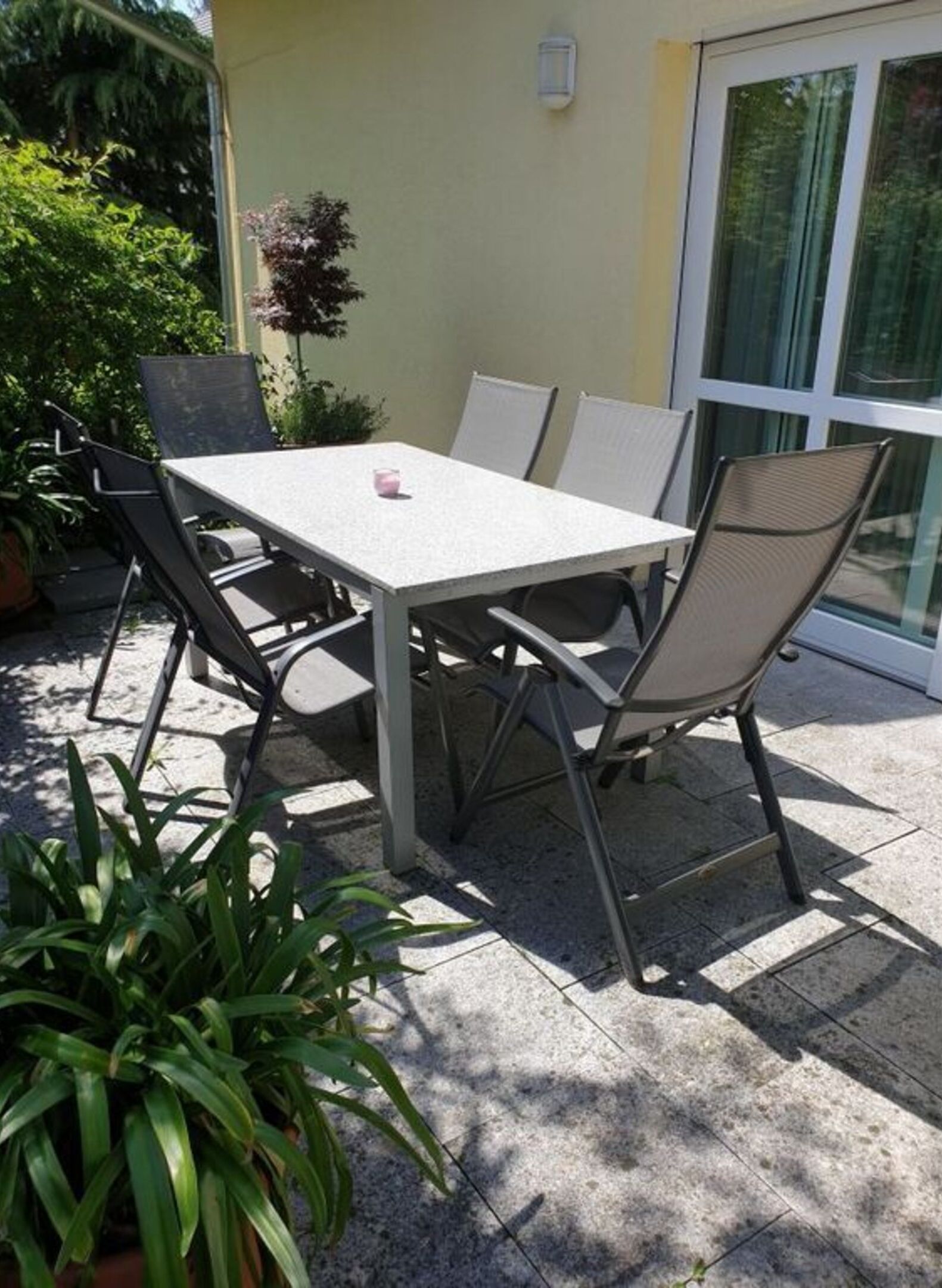 Un patio al aire libre con una mesa y sillas, rodeado de plantas en macetas y un ventanal corredero.