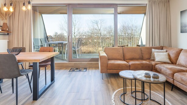 A modern living room with a large sliding glass door leading to a balcony, a brown leather sofa, a wooden dining table with chairs, and a small round coffee table. - Apartment on the 2nd Floor for Family (Middelkerke)