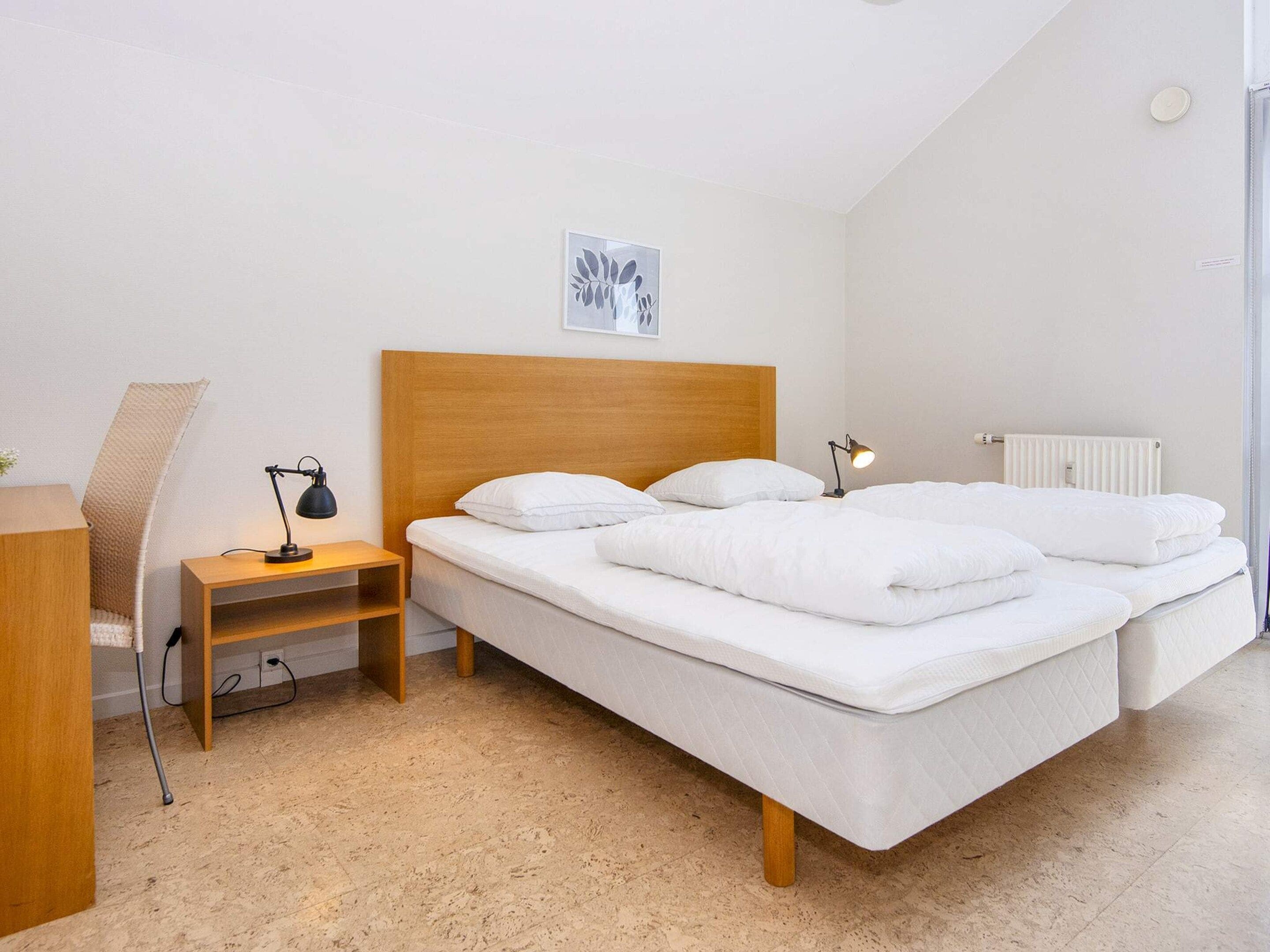 A bedroom with a wooden headboard, a bed with white bedding, a bedside table with a lamp, a radiator, and a framed picture on the wall.