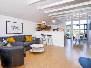 Een moderne woonkamer met een grijze bank, twee ronde tafels en een keukenhoek met een bar en krukken.