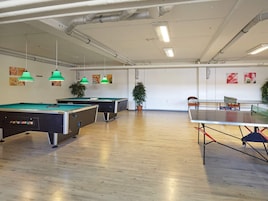 Un área de recreación con mesas de billar y de tenis de mesa, piso de madera y lámparas colgantes verdes.