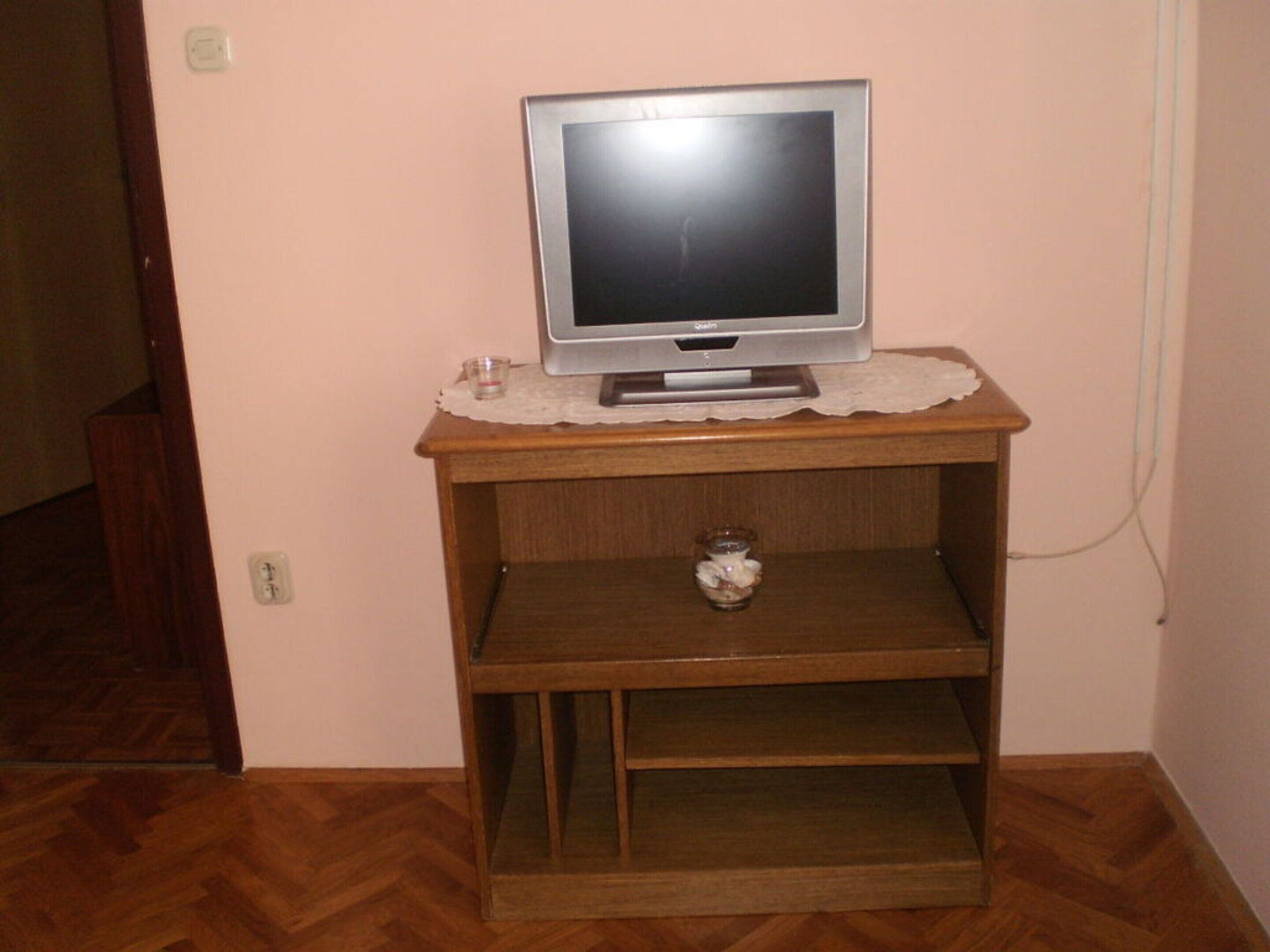 Un televisor sobre un soporte de madera con un copo de encaje, un pequeño recipiente de vidrio y un enchufe en la pared.