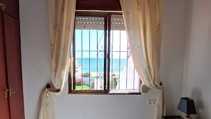Une chambre avec une fenêtre donnant sur la plage, un lit, une table de chevet avec une lampe et une armoire en bois.