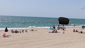 Une plage où des gens se détendent sous des parasols, une grande formation rocheuse et l’océan en arrière-plan.