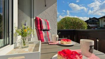 A dining table with a slice of cake and a mug, a vase with flowers, and a checkered napkin on a balcony.