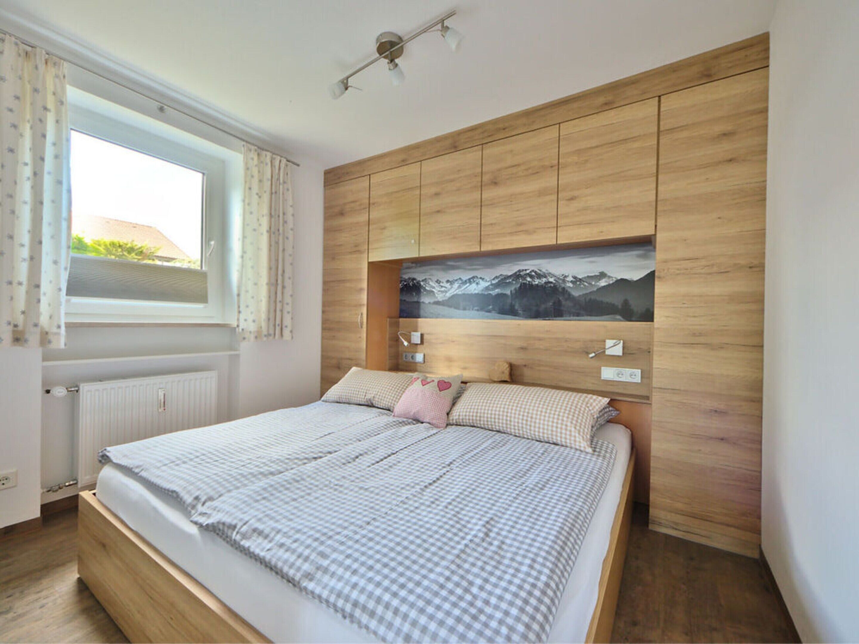 A bedroom with a wooden headboard, a bed with checkered bedding, a window with curtains, and a wall-mounted light fixture.