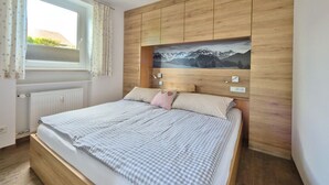 A bedroom with a wooden headboard, a bed with checkered bedding, a window with curtains, and a wall-mounted light fixture. - Pleasant Apartment in Fischen (Fischen im Allgaeu)