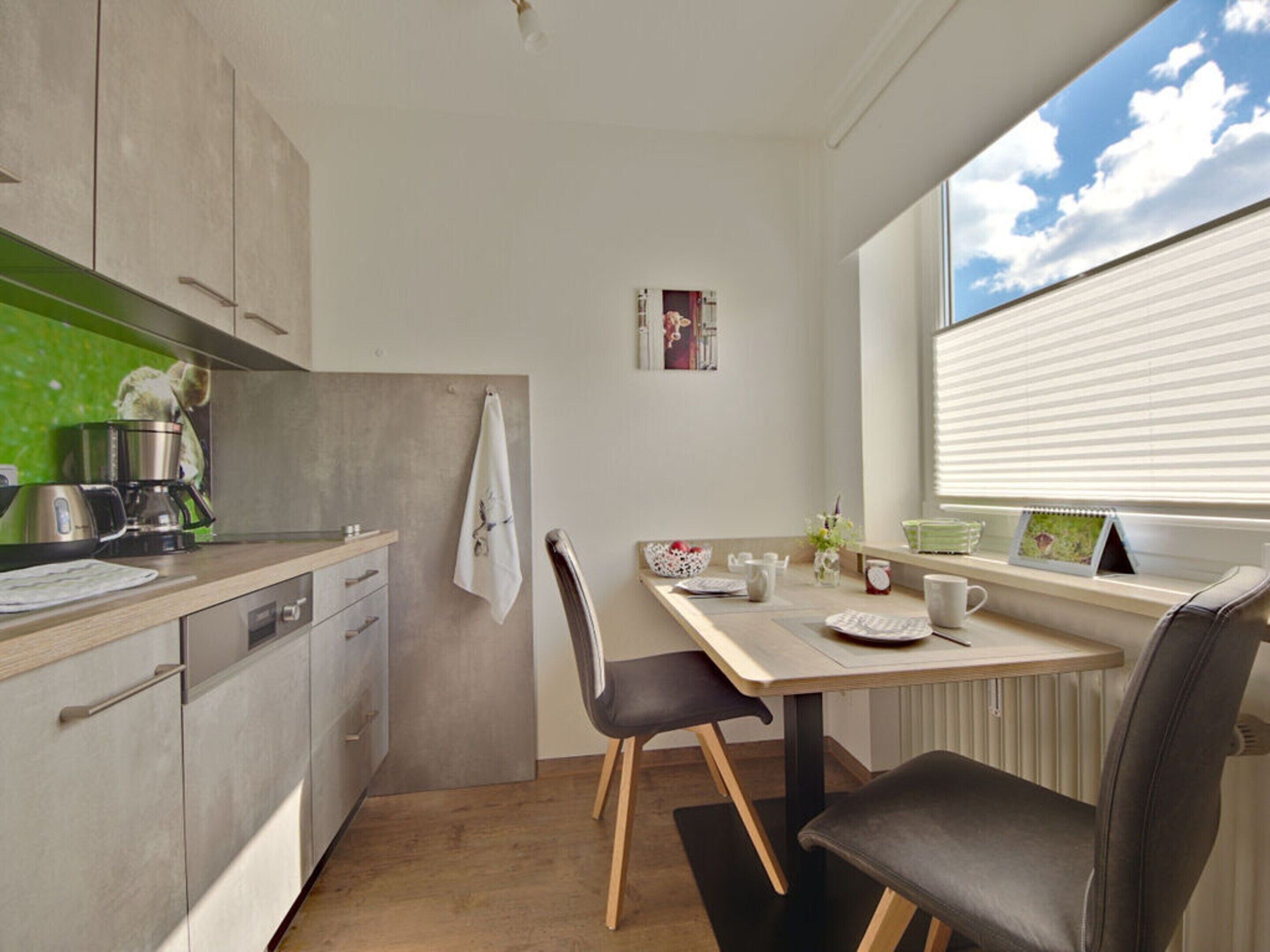 A compact kitchen with a dining area, featuring a window with blinds, a coffee maker, and a small table with chairs.