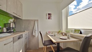 A compact kitchen with a dining area, featuring a window with blinds, a coffee maker, and a small table with chairs. - Pleasant Apartment in Fischen (Fischen im Allgaeu)