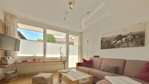 A modern living room with a sofa, ottoman, and coffee table, a TV, and a window with a view of the outside. - Pleasant Apartment in Fischen (Fischen im Allgaeu)