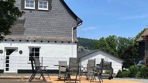 A white house with a grey roof, a wooden deck with outdoor furniture, and a fenced garden area.