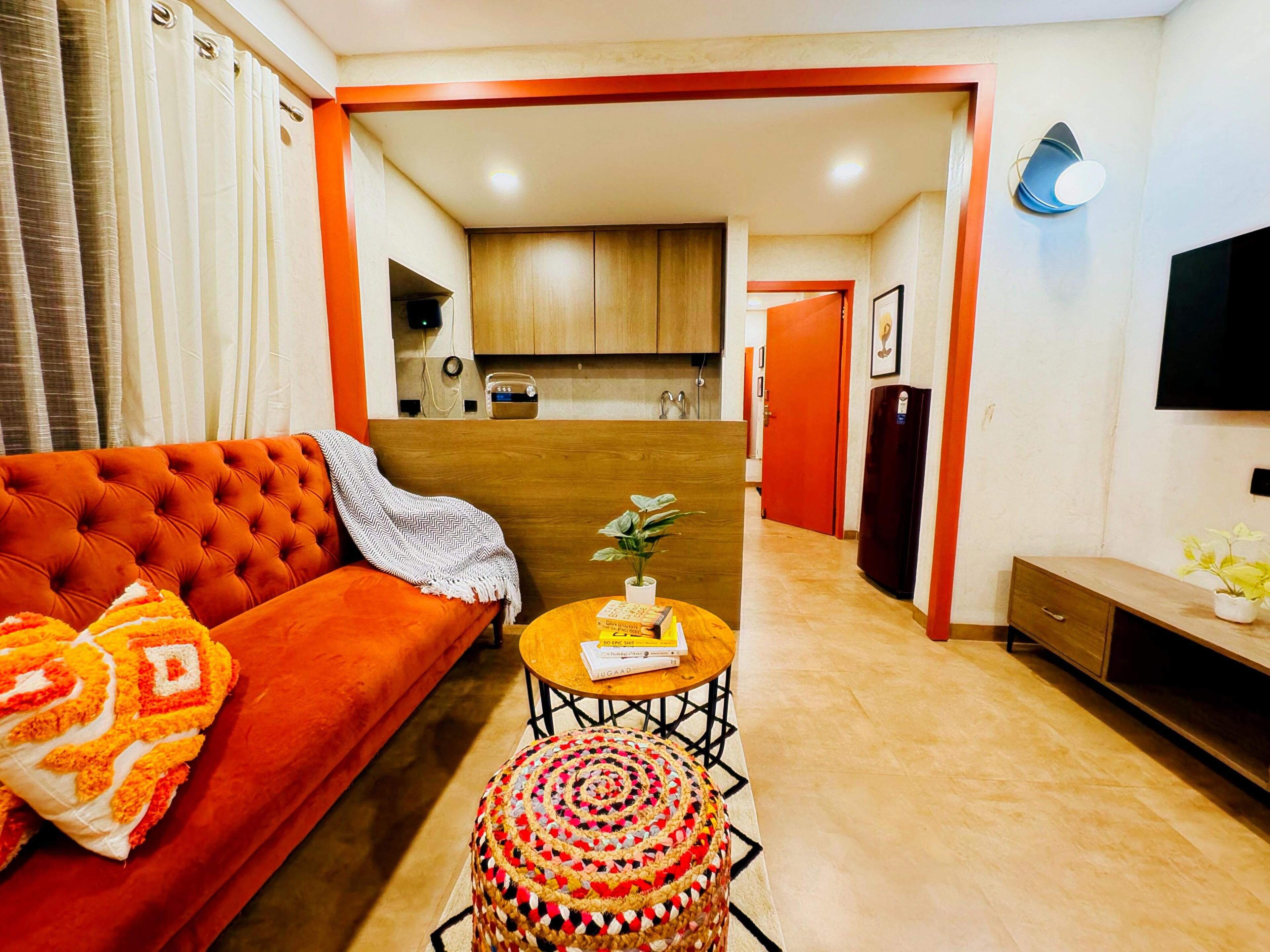 Una sala de estar con sofá, reposapiés y mesa de centro. Se ve una cocina al fondo con armarios y un refrigerador.