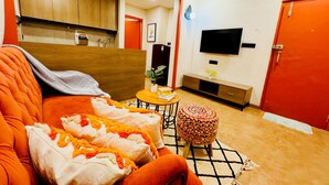 Un salon moderne avec un canapé, une petite table ronde, un téléviseur et, au fond, un coin cuisine.