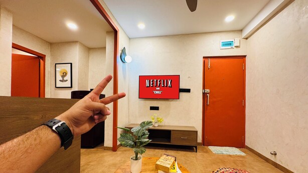 Un hall d’hôtel avec une table ronde en bois, une plante en pot, une bibliothèque, un téléviseur fixé au mur qui affiche « NETFLIX AND CHILL », et une porte rouge.