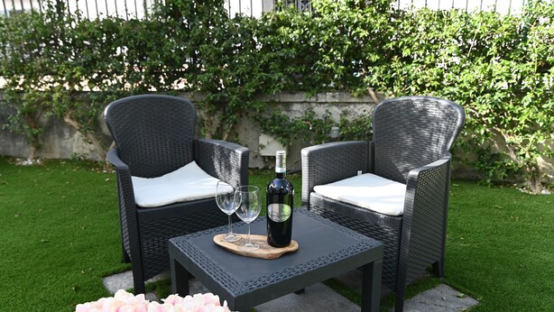 Deux fauteuils en osier avec des coussins blancs, une petite table sur laquelle se trouvent une bouteille de vin et deux verres, et un bouquet de fleurs roses.