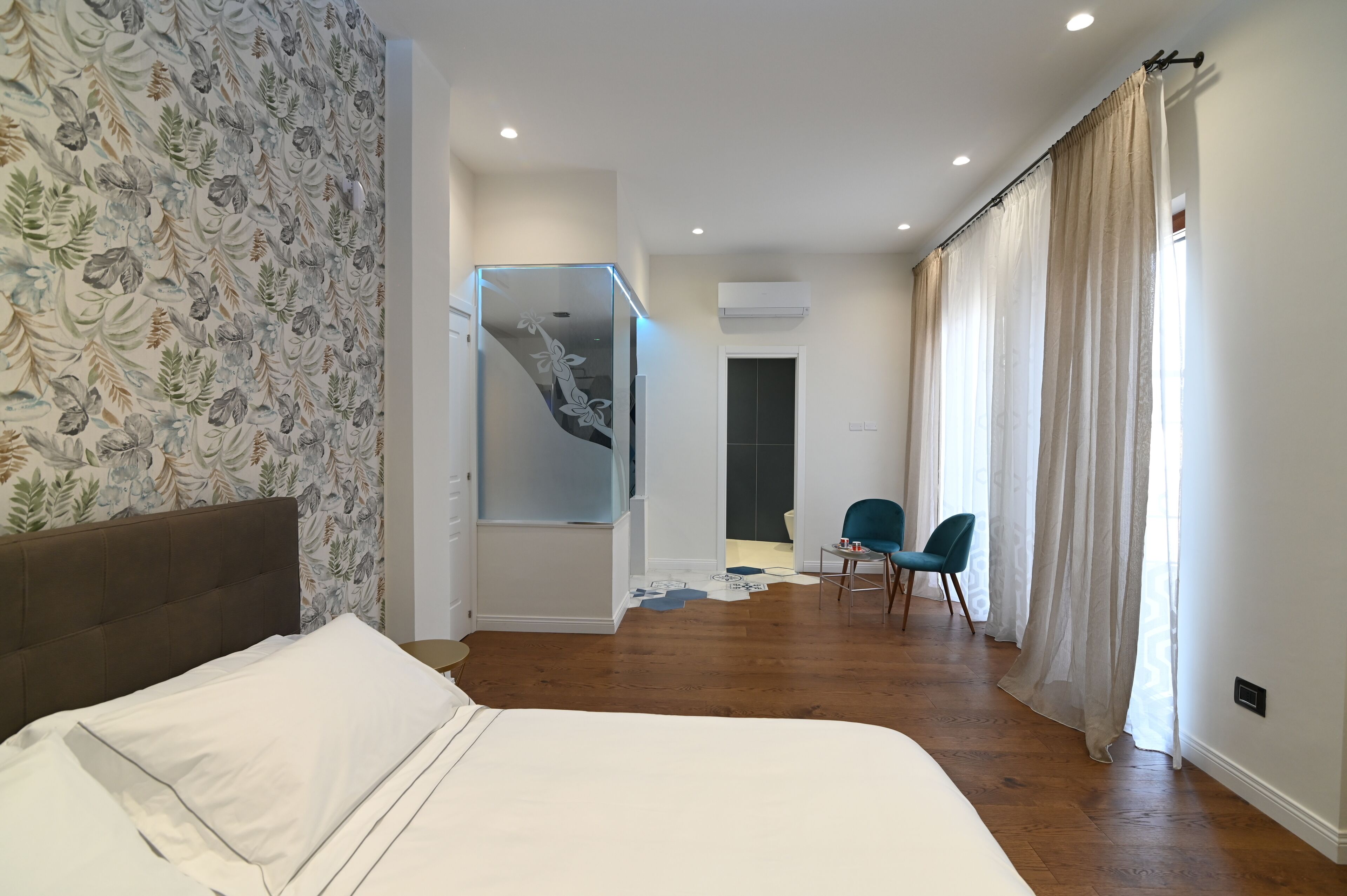 A bedroom with a bed, a chair, a desk, and a bathroom visible through a glass door.
