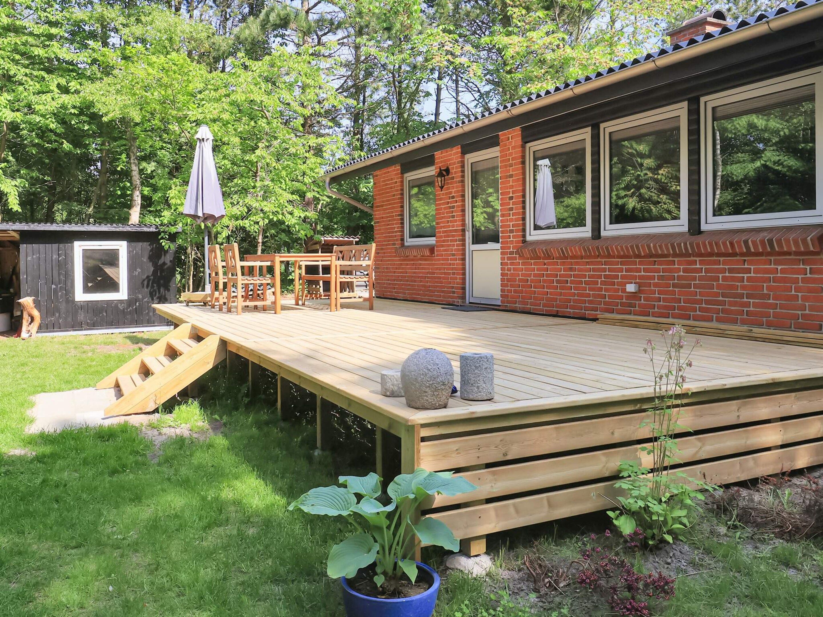 A wooden deck with a table and chairs, a small shed, and a house with a brick exterior and large windows.