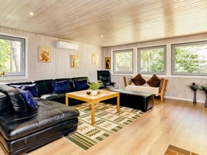 A modern living room with a black leather sofa, a wooden coffee table, and a black leather armchair. There are large windows providing a view of greenery outside. - 8 Person Holiday Home in Hadsund (Hadsund)