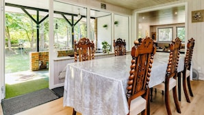 Un comedor con una mesa larga cubierta por un mantel blanco, rodeada de sillas de madera con intrincados grabados. La habitación tiene ventanas amplias que dejan entrar luz natural y ofrecen vistas a un jardín.