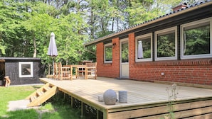Una terraza de madera con un pequeño espacio de estar al aire libre, una casa de ladrillo y un cobertizo en un jardín.