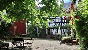 Een geplaveid binnenhof met terras, omgeven door weelderige begroeiing en een traditioneel vakwerkhuiz.