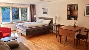 Een slaapkamer met een bed, nachtkastje, lamp en een klein bureau met stoel. Tegen de muur staat een boekenkast en er is een raam met gordijnen.