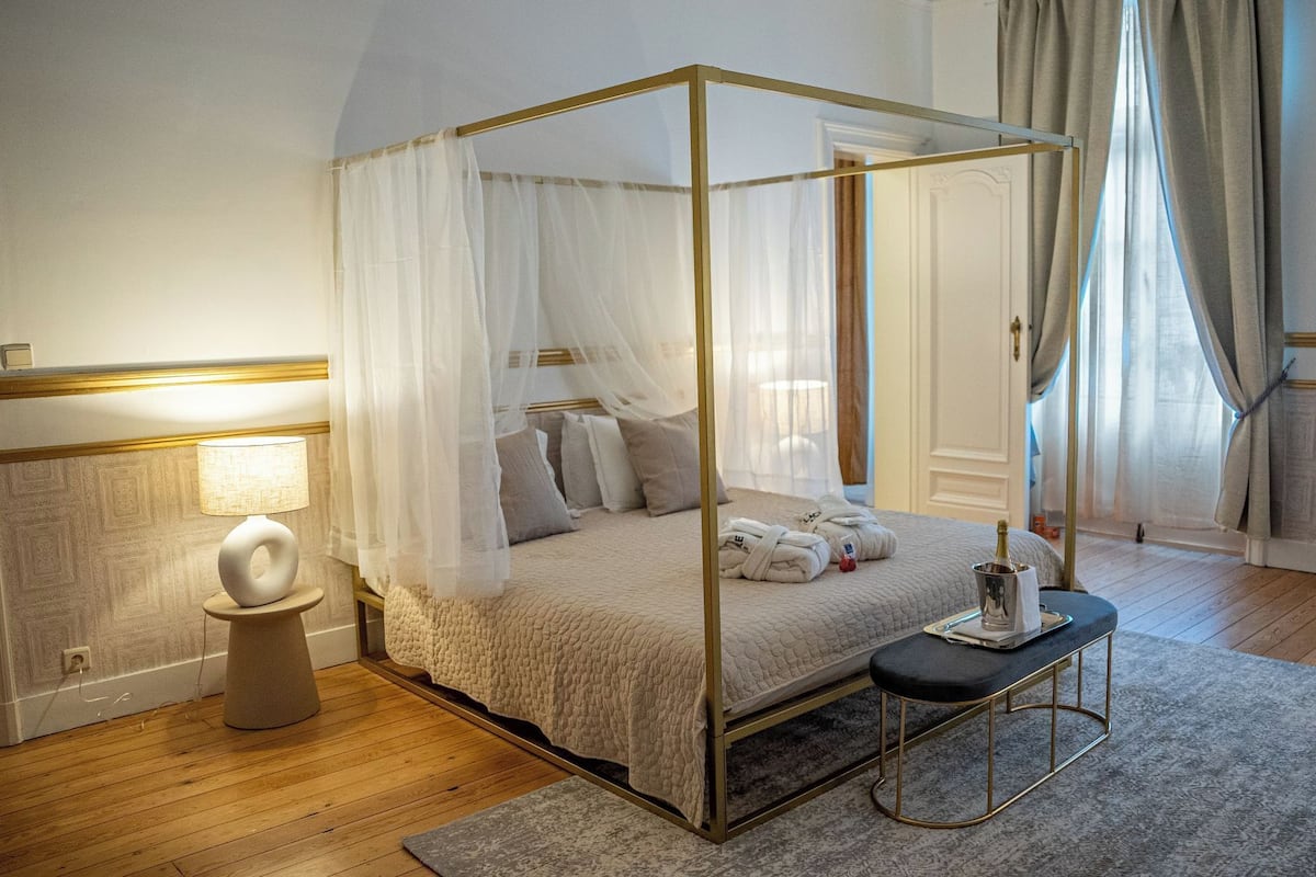 Ein Himmelsbett mit Baldachin, ein Nachttisch mit Lampe, eine Bank und eine Champagnerflasche.