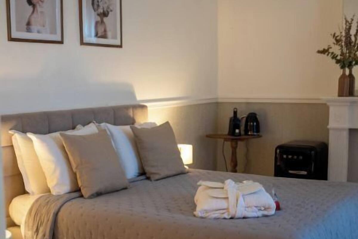 Ein ordentlich bezogenes Bett mit beiger Decke und Kissen, ein kleiner Tisch mit Wasserkocher und Blumenvase sowie ein Spiegel an der Wand.