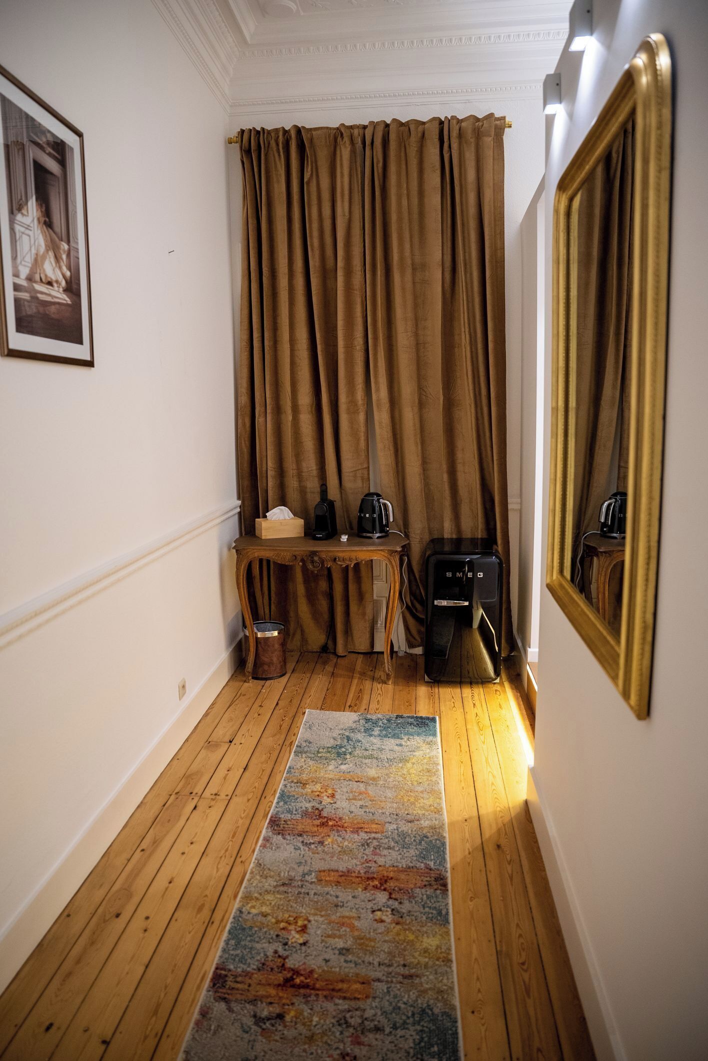 A room with a wooden floor, a patterned rug, a desk with a coffee maker, a trash bin, and a large mirror.
