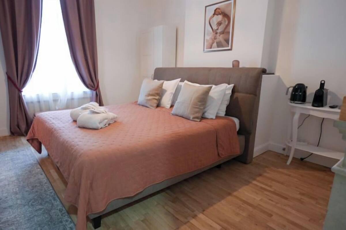 Ein ordentlich bezogenes Bett mit einer rosa Tagesdecke und weißen Kissen, ein kleiner Nachttisch mit einer Kaffeemaschine und ein Gemälde an der Wand.