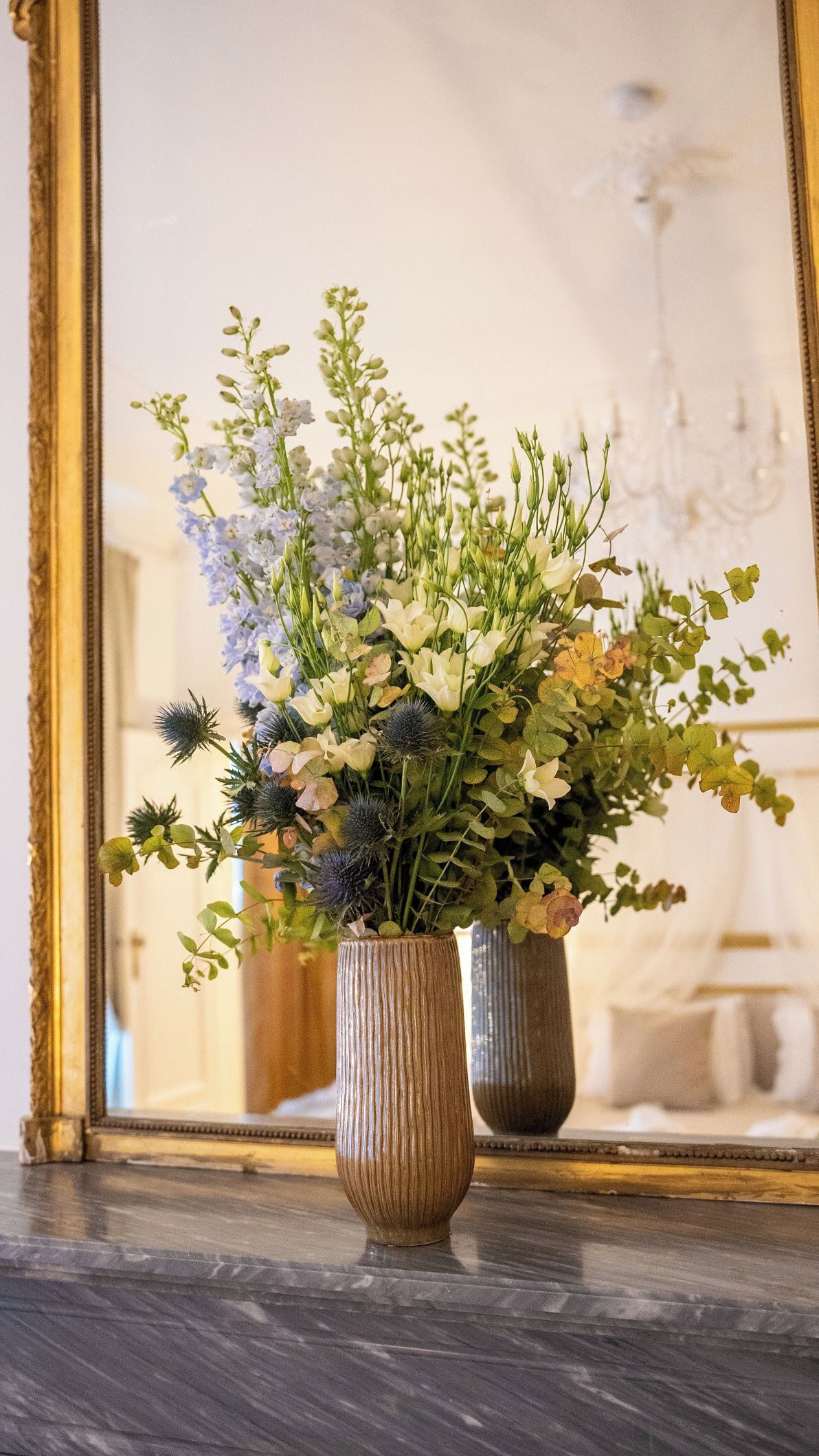 Two vases with flowers on a marble surface, a mirror reflecting a chandelier and a room interior.