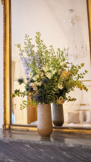 Deux vases ornés de fleurs sont posés sur une surface en marbre, un miroir reflète un lustre et l’intérieur d’une pièce.