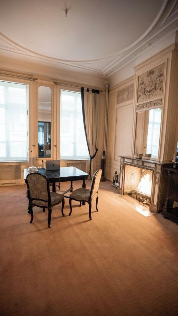 Ein Zimmer mit einem runden Tisch, zwei Stühlen, einem Kamin und großen Fenstern mit Vorhängen.