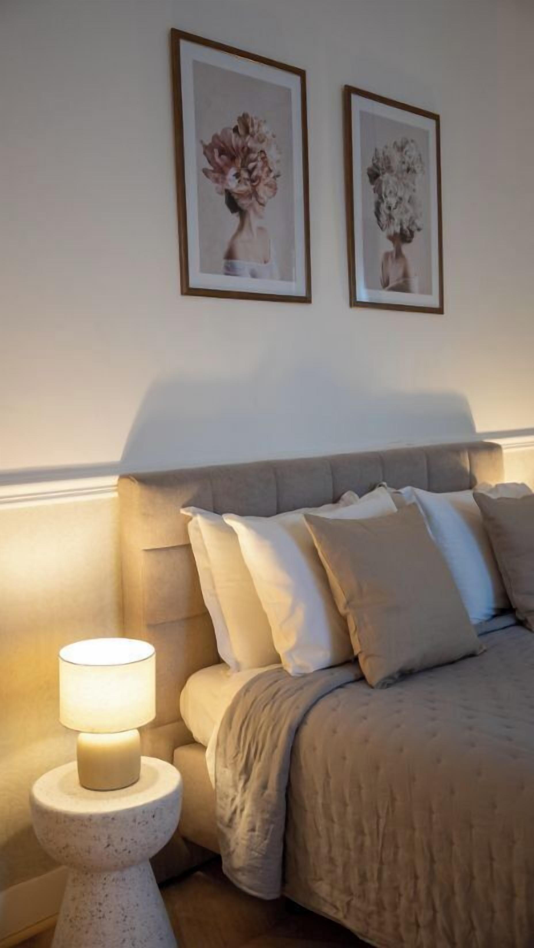 Een slaapkamer met een bed, kussens, een nachtkastje met een lamp en twee kunstwerken in lijsten aan de muur.