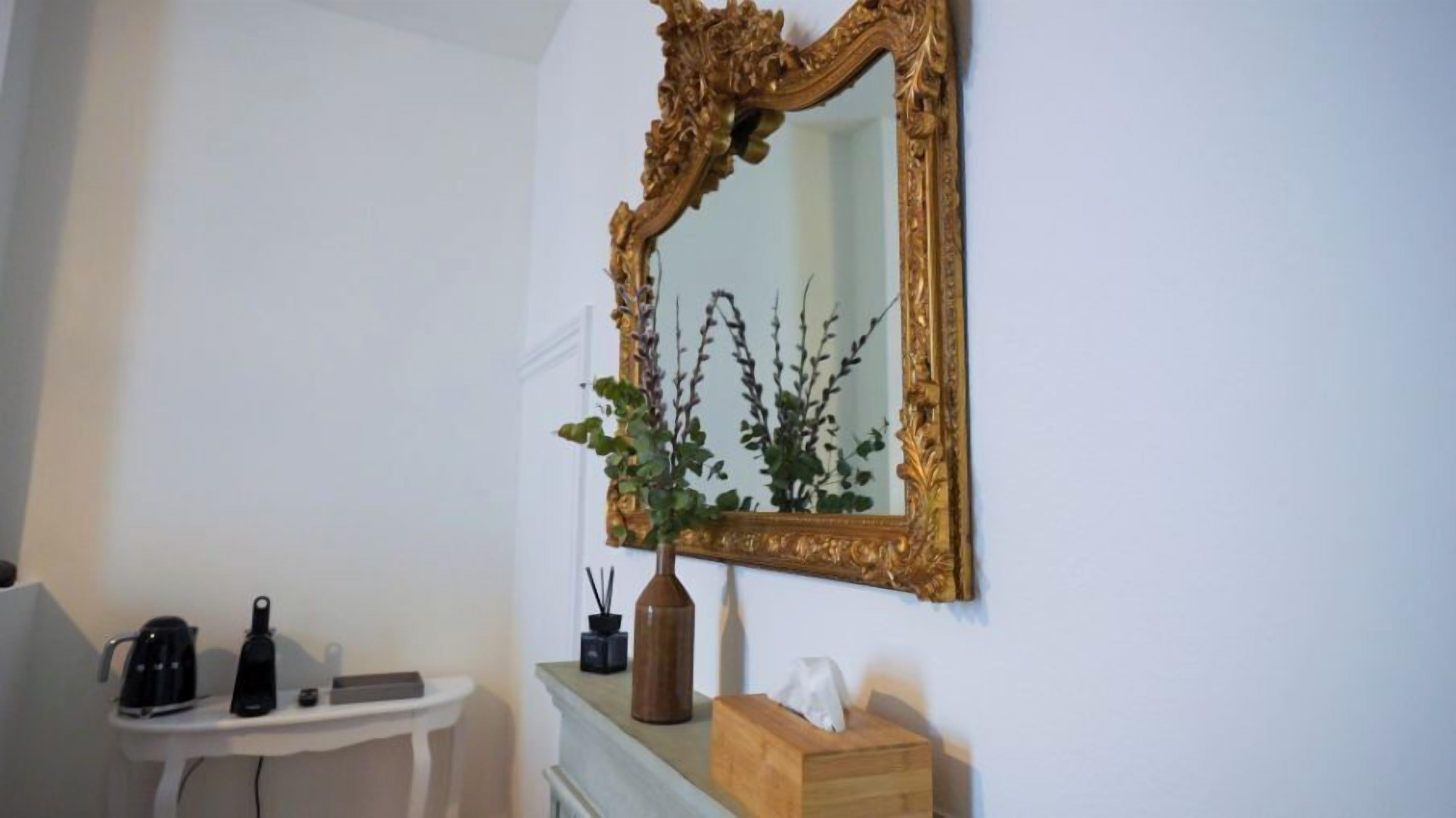 Een badkamer met een wit wastafelmeubel, een spiegel met gouden lijst, een potplant en toiletartikelen.