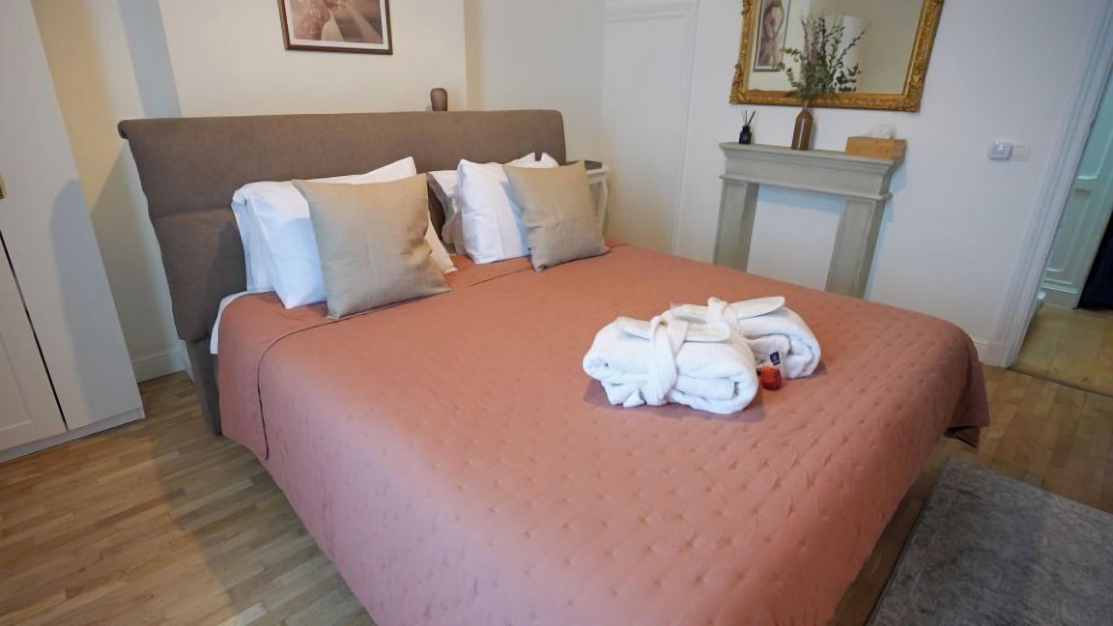 Een net opgemaakt bed met een roze sprei en witte kussens, een houten vloer en een klein wit tafeltje met een vaas en een spiegel erboven.