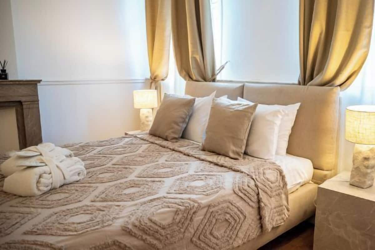 Ein ordentlich bezogenes Bett mit einer gemusterten Tagesdecke, zwei Nachttischlampen und einem Nachttisch mit Lampe.