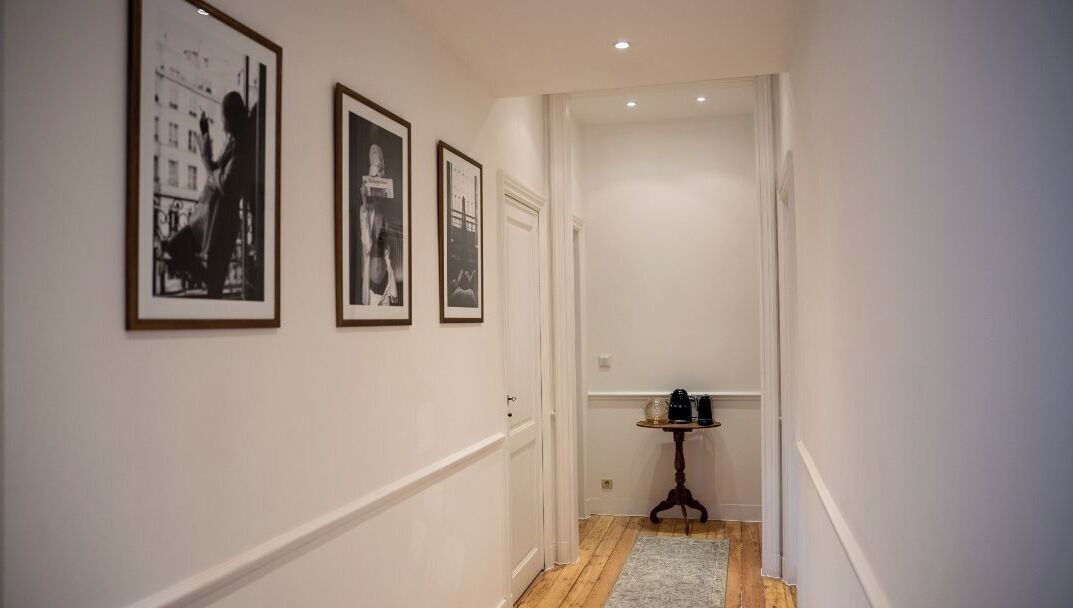 A hallway with framed black and white photographs on the wall, a small round table with a lamp, and a door at the end.
